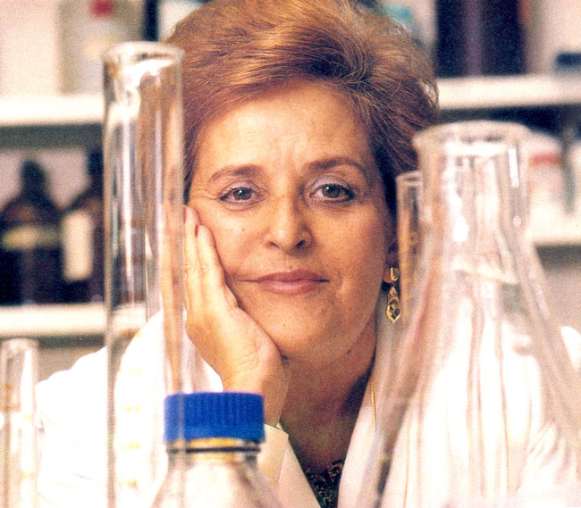 Una mujer se apoya con la mano en la cara en una encimera de un laboratorio. Se la ve entre dos matraces que están cerca de la cámara